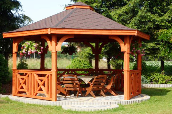 Šesťstranný drevený altánok MONTNA vo farbe teak s jednym vstupom, zábradlim a dreveným záhradným nábytkom stojací v záhrade